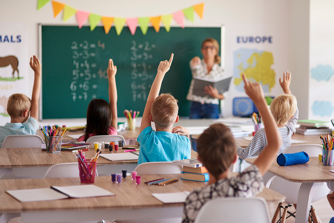 Elementary school students raising hands during math lesson in classroom with teacher at chalkboard